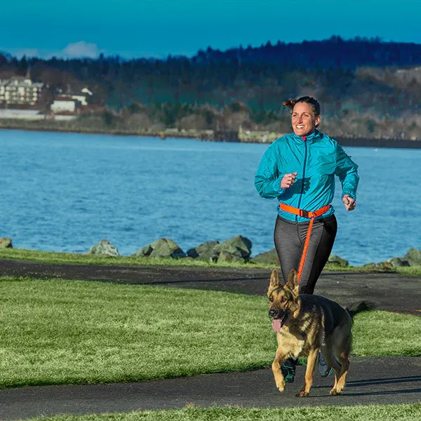 A person running on a concrete pathway next to grass and and river, using a FurHaven Trail Pup Hands-Free Leash to run with their German Shepherd