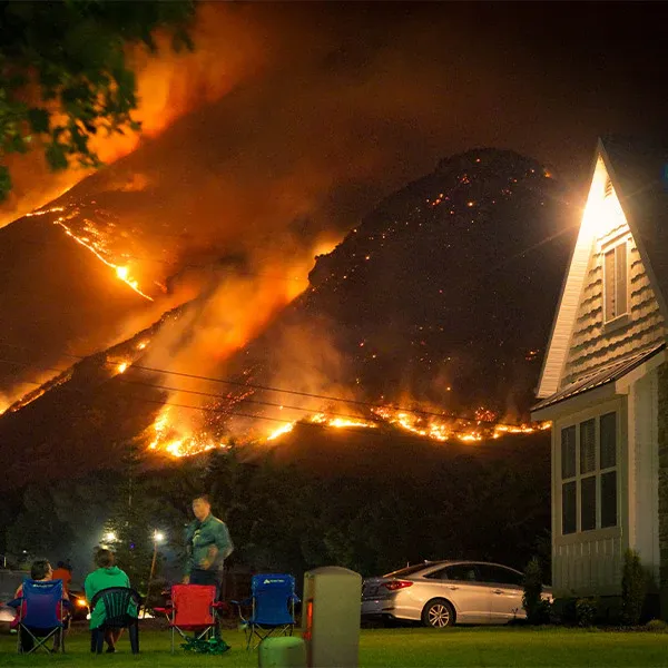 A wildfire rages on a hill while several people sit in camp chairs near their house and observe the phenomenon, at FurHaven Pet Products